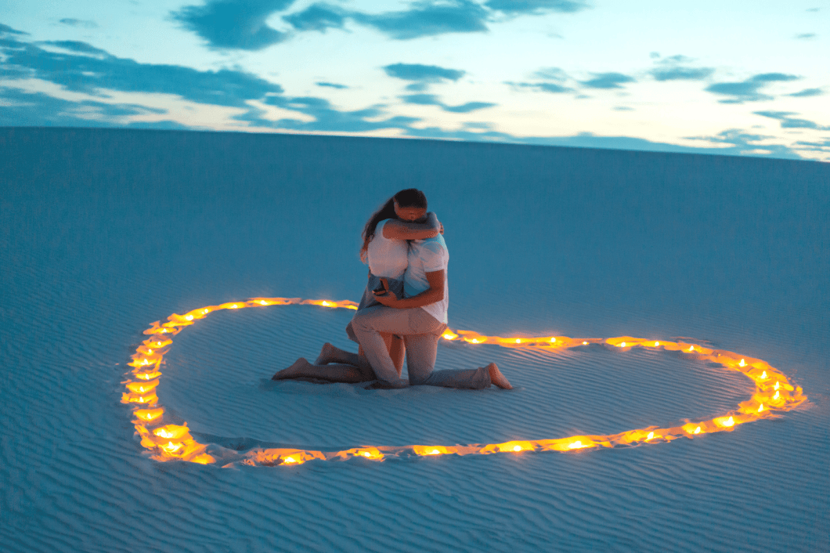Man and woman kneeling and hugging among heart-shaped lights on sand